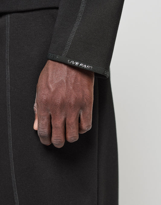 A close-up of a hand resting on black pants, wearing the LIVE FAST DIE YOUNG LFDY Active Fleece Hooded Zip in an Oversize Fit with LIVE FAST text on the sleeve cuff.