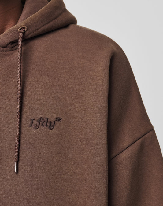 Close-up of a person wearing the LIVE FAST DIE YOUNG Hooded Zip with the hood up. The brown hoodie showcases subtle Lfdj™ embroidery on the chest in a darker shade, with drawstrings visible against a plain background.
