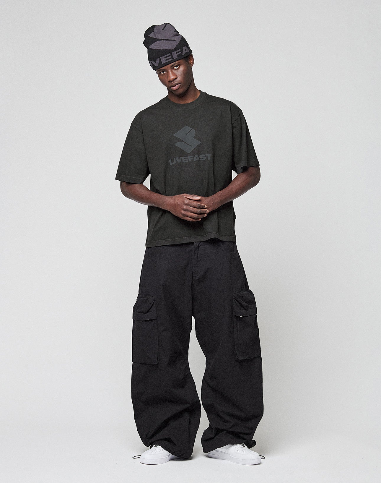 A person wearing a dark beanie, a loose LIVE FAST DIE YOUNG T-shirt with “LIVEFAST” on the front, LFDY Baggy Cargo pants in 100% cotton, and white sneakers stands against a plain light background.