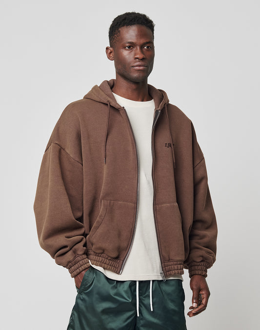 A man stands against a plain background wearing the LIVE FAST DIE YOUNG Hooded Zip over a white textured shirt and dark green satin pants, hands in his pockets, with a neutral facial expression.