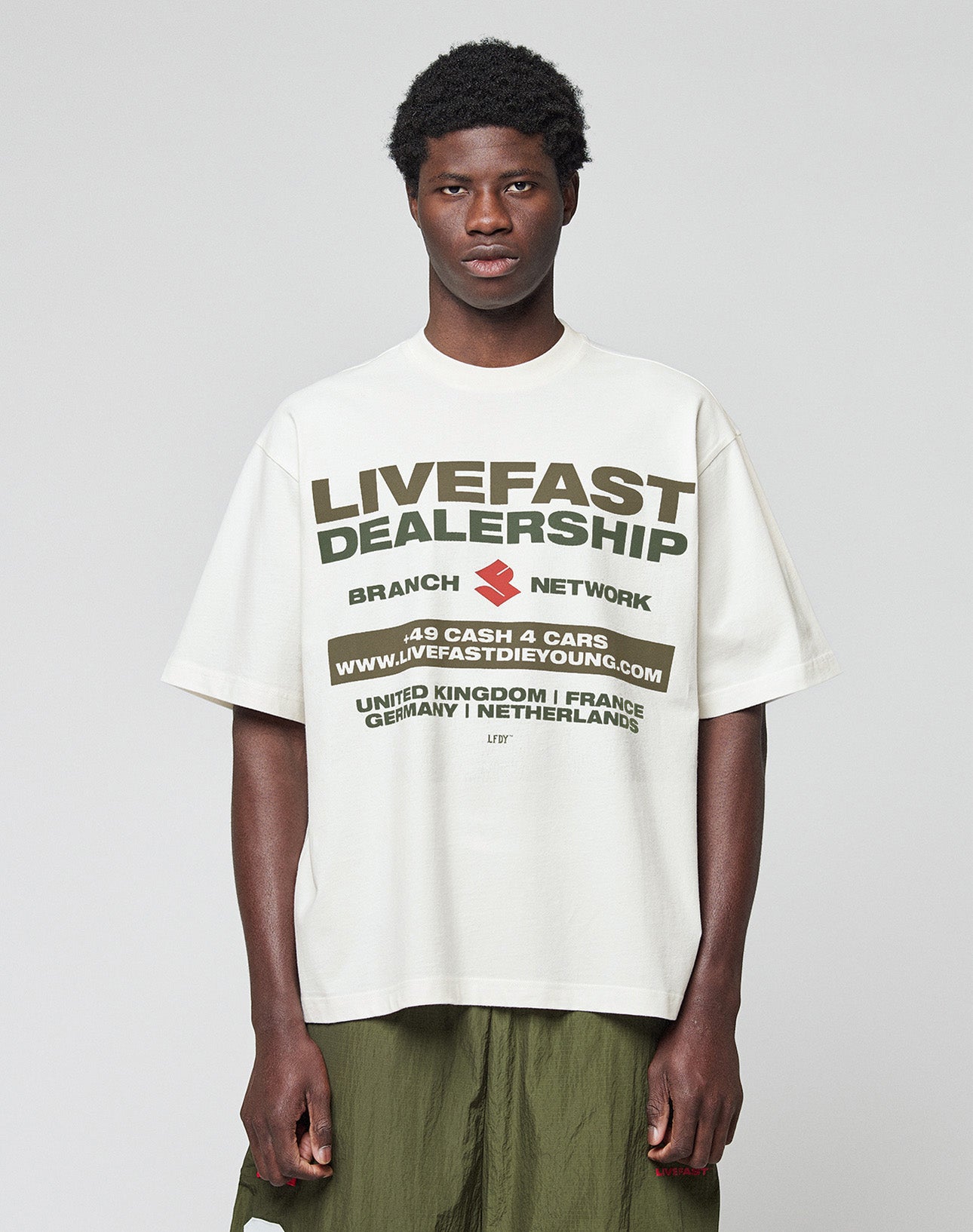 A young man with short curly hair wears the LFDY Network Tee by LIVE FAST DIE YOUNG, an oversized white 100% Baumwolle T-shirt with a bold black and olive green dealership-inspired front print, standing against a light gray background.