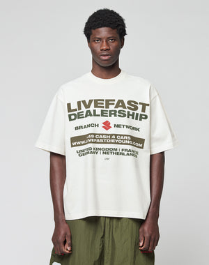 A young man with short curly hair wears the LFDY Network Tee by LIVE FAST DIE YOUNG, an oversized white 100% Baumwolle T-shirt with a bold black and olive green dealership-inspired front print, standing against a light gray background.