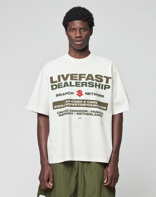 A young man with short curly hair wears the LFDY Network Tee by LIVE FAST DIE YOUNG, an oversized white 100% Baumwolle T-shirt with a bold black and olive green dealership-inspired front print, standing against a light gray background.