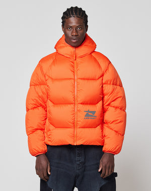 A man faces forward in the LIVE FAST DIE YOUNG Live Fast Puffer Jacket, an orange oversized style with a high collar and reflective "LIVE FAST" print plus graphic, paired with dark pants against a plain light background.