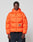 A man faces forward in the LIVE FAST DIE YOUNG Live Fast Puffer Jacket, an orange oversized style with a high collar and reflective "LIVE FAST" print plus graphic, paired with dark pants against a plain light background.