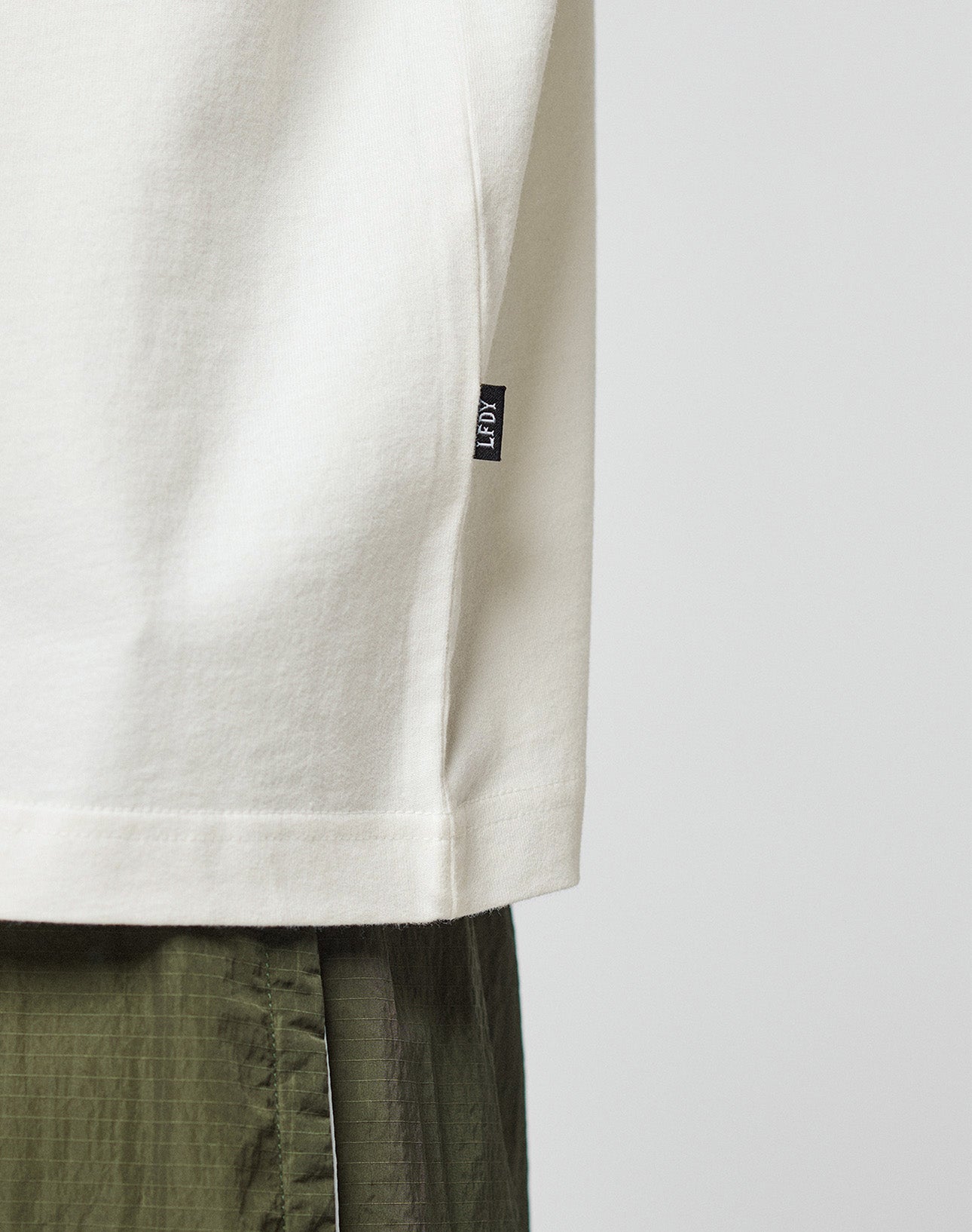 A close-up side view of a person wearing the LIVE FAST DIE YOUNG LFDY Network Tee in off-white with a small black tag, paired with olive green pants, against a plain light background.