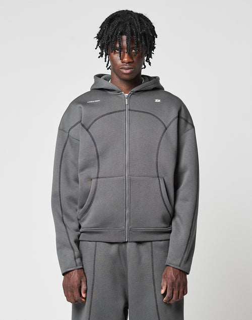 A person with medium-length curly hair wears the LFDY Active Fleece Hooded Zip by LIVE FAST DIE YOUNG in grey Oversize Fit, paired with matching pants, standing against a plain light background and looking directly at the camera with a neutral expression.