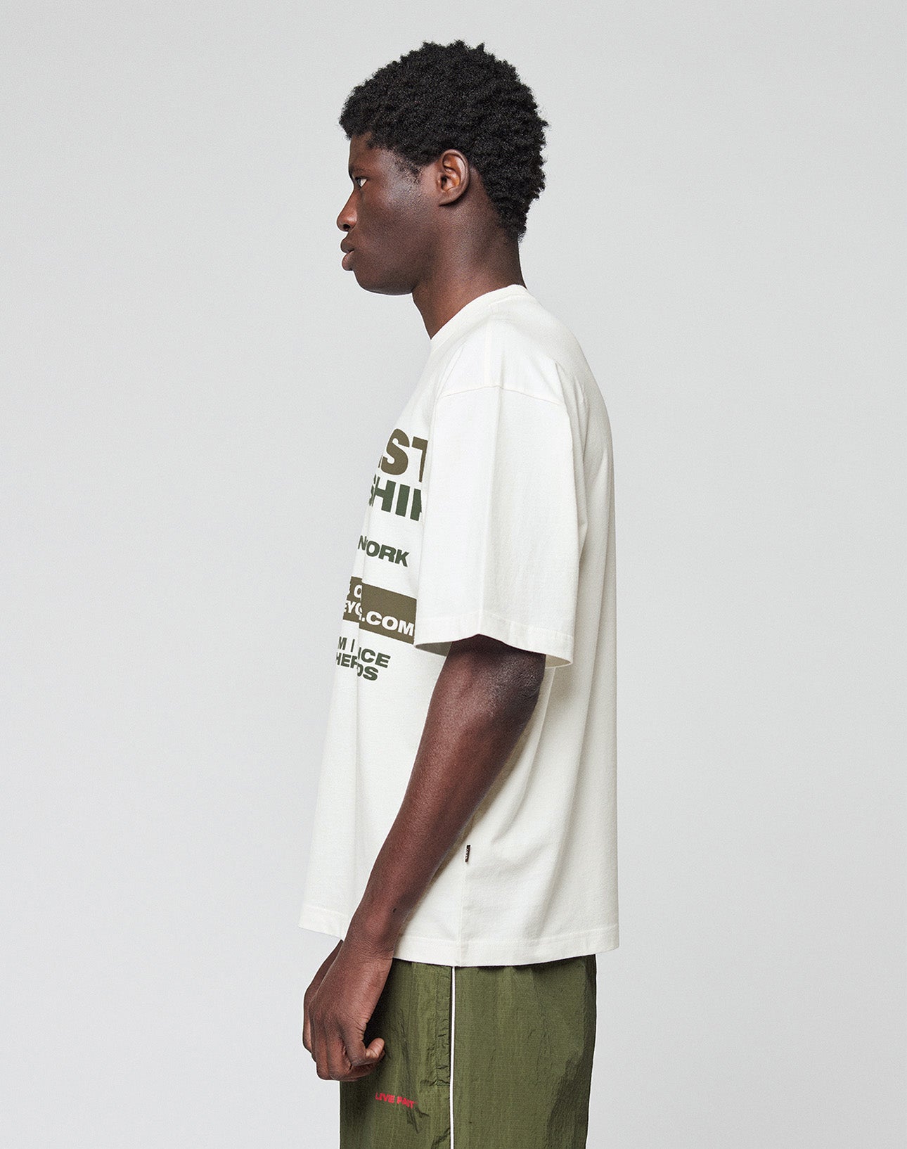 A person with short curly hair and a neutral expression stands in profile against a plain background, wearing the LFDY Network Tee by LIVE FAST DIE YOUNG—an oversize white graphic t-shirt with green frontprint text—and olive shorts.