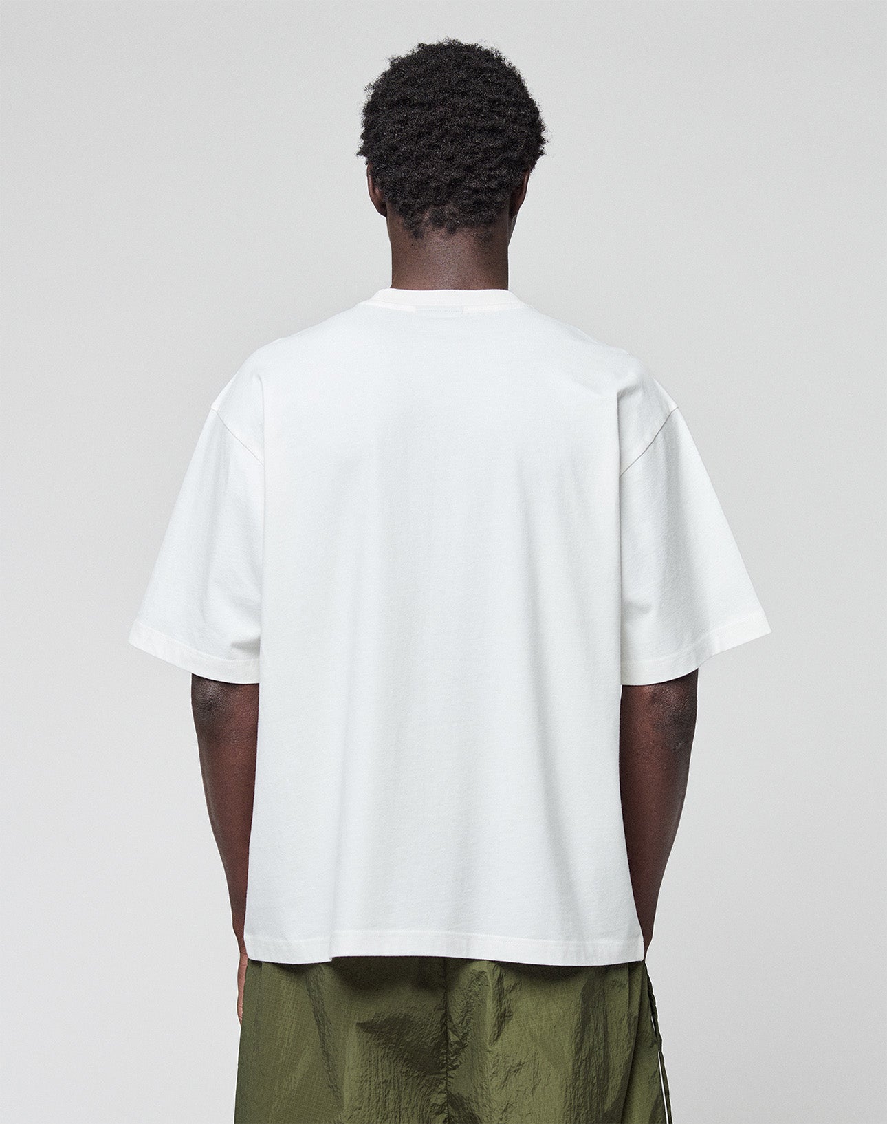 A person with short, curly hair stands with their back to the camera, wearing a white LIVE FAST DIE YOUNG LFDY Network Tee in an oversize fit and olive green pants against a plain, light background.