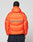 A person with braided hair wears the Live Fast Puffer Jacket by LIVE FAST DIE YOUNG—an oversize, bright orange jacket with bold blue LIVE FAST lettering and a reflektierender Print—standing against a plain, light background.