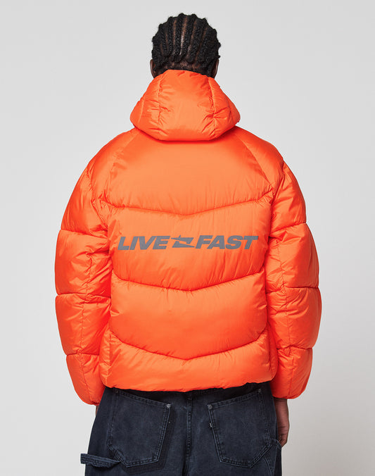 A person with braided hair wears the Live Fast Puffer Jacket by LIVE FAST DIE YOUNG—an oversize, bright orange jacket with bold blue LIVE FAST lettering and a reflektierender Print—standing against a plain, light background.