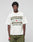 A person with a neutral expression stands against a gray background, wearing the LIVE FAST DIE YOUNG LFDY Network Tee—an oversized, 100% cotton white T-shirt with bold front print—and olive green pants.