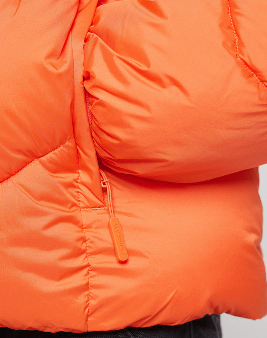 Close-up of the LIVE FAST DIE YOUNG Live Fast Puffer Jacket in orange, featuring a zippered pocket with a matching pull, shiny quilted fabric with visible stitching, slight puffiness, and a reflektierender Print for added visibility.