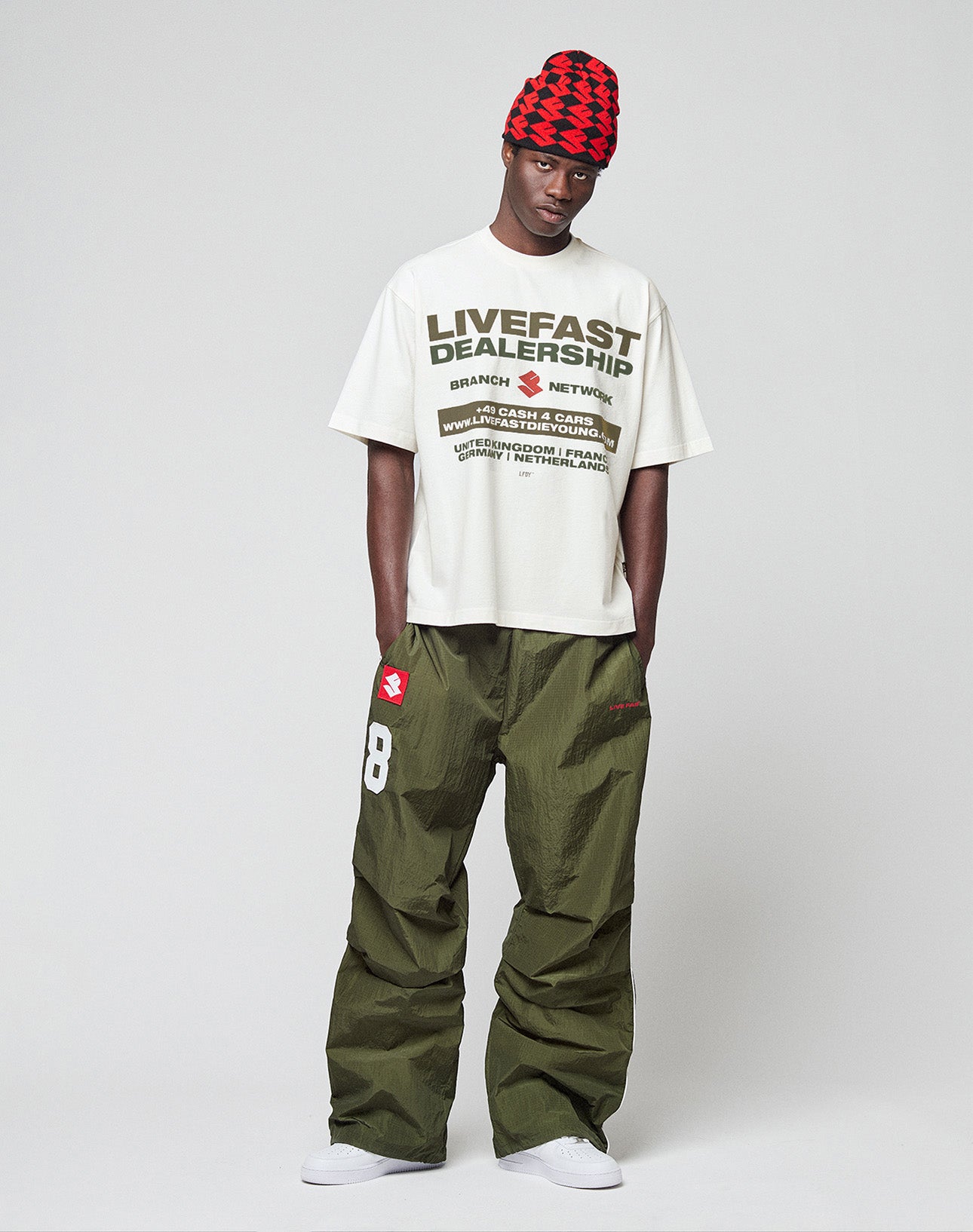 A person stands against a plain background wearing the LFDY Network Tee by LIVE FAST DIE YOUNG, loose olive green pants with a number 8 and red patch, white sneakers, and a red and black patterned beanie.