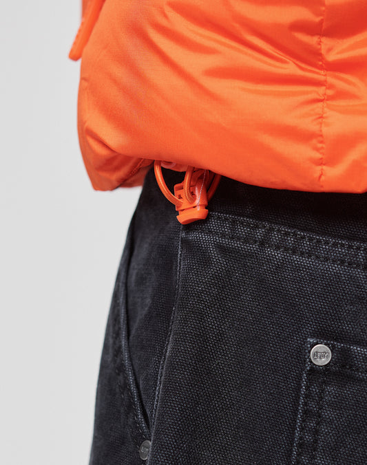 Close-up of the LIVE FAST DIE YOUNG Live Fast Puffer Jacket in orange, featuring an oversize fit and water-repellent fabric, partially covering dark pants with an orange plastic cord lock at the hem.