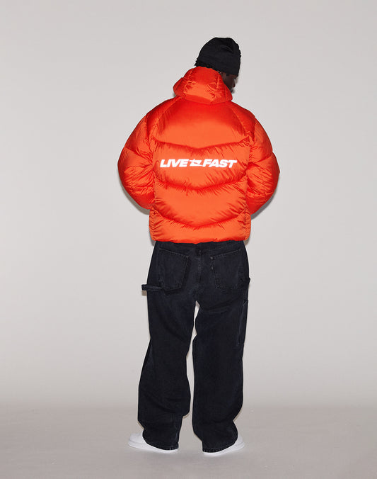 A person faces away, wearing the LIVE FAST DIE YOUNG Live Fast Puffer Jacket in bright orange with reflective "LIVE FAST" print on the back, paired with black pants, a black beanie, and white shoes against a plain light background.
