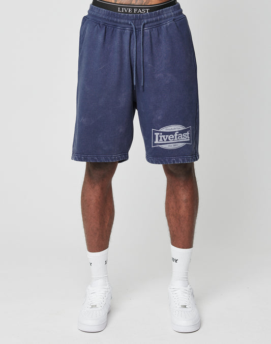 A person wearing LIVE FAST DIE YOUNG LFDY Bulge Shorts in navy blue with drawstring and vintage wash, paired with white socks and sneakers, stands against a plain light background. Only the lower body is visible.