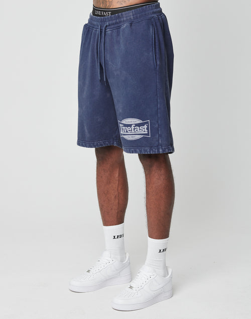 A person in blue LFDY Bulge Shorts by LIVE FAST DIE YOUNG with vintage wash, elastic waistband, drawstring, and logo near the hem, paired with white Live Fast socks and white Nike Air Force 1s against a plain white background.