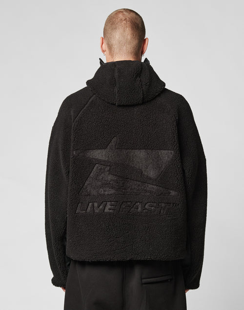 Seen from behind, a person with short hair wears the oversized black Live Fast Teddy Jacket by LIVE FAST DIE YOUNG, featuring embossed branding, a large graphic, a visor-hood opening, and the words LIVE FAST on textured fabric. The background is light-colored.