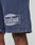 Close-up of a person’s leg wearing LIVE FAST DIE YOUNG LFDY Bulge Shorts in blue with vintage wash, elastic waistband, “Everybody Eats” patch above a large livefast logo, and “Est. 2013” print. Made from 100% cotton.