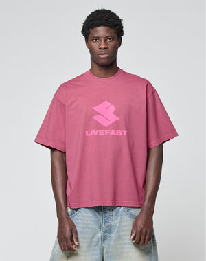 A person wears the Live Fast LF Tee by LIVE FAST DIE YOUNG—an oversize pink T-shirt with a bold "LIVEFAST" frontprint and stylized logo, made from 100% Baumwolle—paired with light blue jeans against a plain background.