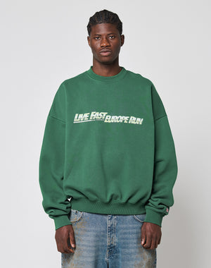 A man in a green LIVE FAST DIE YOUNG LFDY Europe Run Sweater with LIVE FAST EUROPEAN on the front stands facing forward. Made from soft Baumwolle, he pairs it with blue jeans against a light gray background.