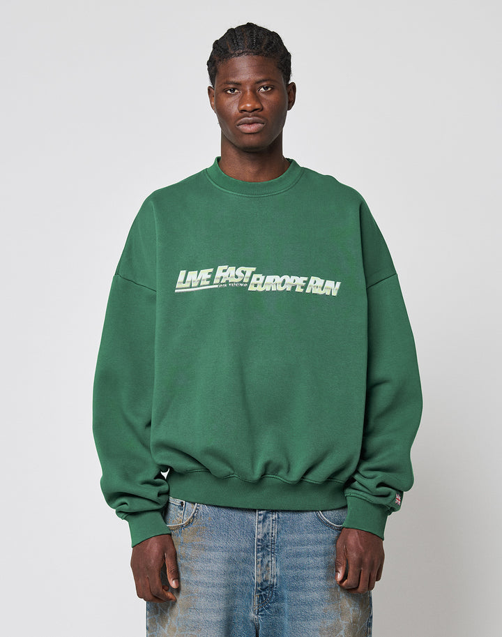 A man in a green LIVE FAST DIE YOUNG LFDY Europe Run Sweater with LIVE FAST EUROPEAN on the front stands facing forward. Made from soft Baumwolle, he pairs it with blue jeans against a light gray background.