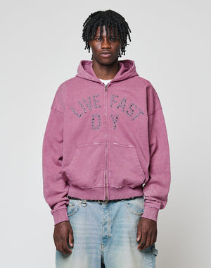 A young man with short dreadlocks wears the Live Fast Rivet Hooded Zip by LIVE FAST DIE YOUNG—a light purple hoodie featuring studded lettering and a handmade spray effect—paired with faded blue jeans, against a plain light gray background.