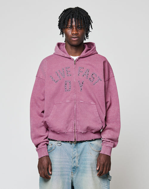 A young man with short dreadlocks wears the Live Fast Rivet Hooded Zip by LIVE FAST DIE YOUNG—a light purple hoodie featuring studded lettering and a handmade spray effect—paired with faded blue jeans, against a plain light gray background.