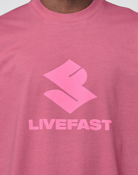 A close-up of a person wearing the Live Fast LF Tee by LIVE FAST DIE YOUNG, featuring a bold abstract front logo above "LIVEFAST" in pink block letters. The oversize fit adds comfort.