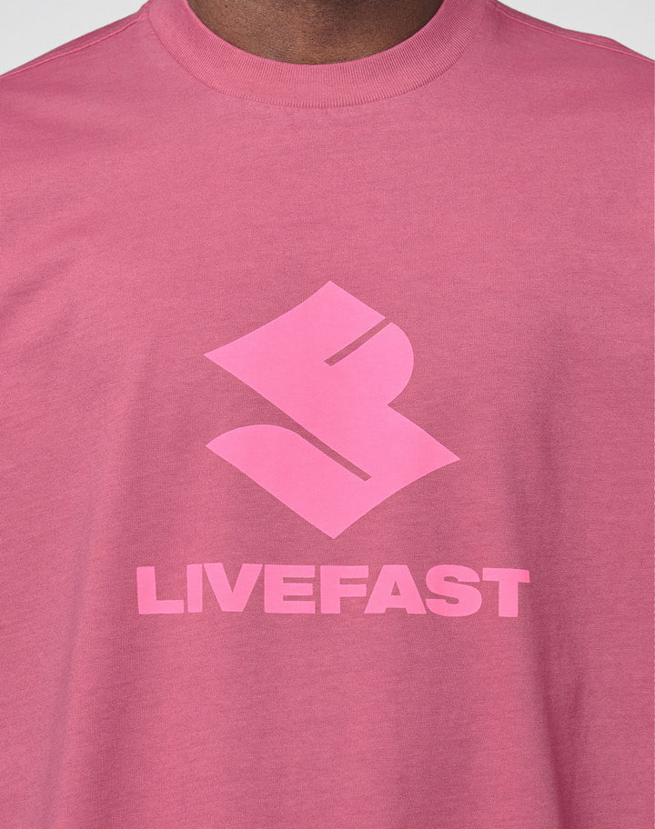 A close-up of a person wearing the Live Fast LF Tee by LIVE FAST DIE YOUNG, featuring a bold abstract front logo above "LIVEFAST" in pink block letters. The oversize fit adds comfort.