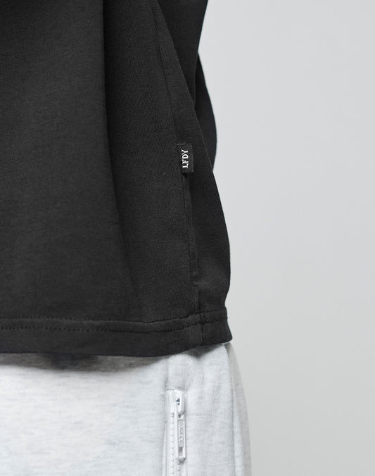 Close-up of a person wearing the LIVE FAST DIE YOUNG Live Fast Paris Tee in black with a small REP tag, made from 100% Baumwolle, paired with light gray pants featuring a white zipper pocket. Plain light background.