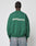 A person with braided hair, facing away, wears the LIVE FAST DIE YOUNG LFDY Europe Run Sweater in green with bold white LIVE FAST RACE AM text on the back, paired with light blue jeans.