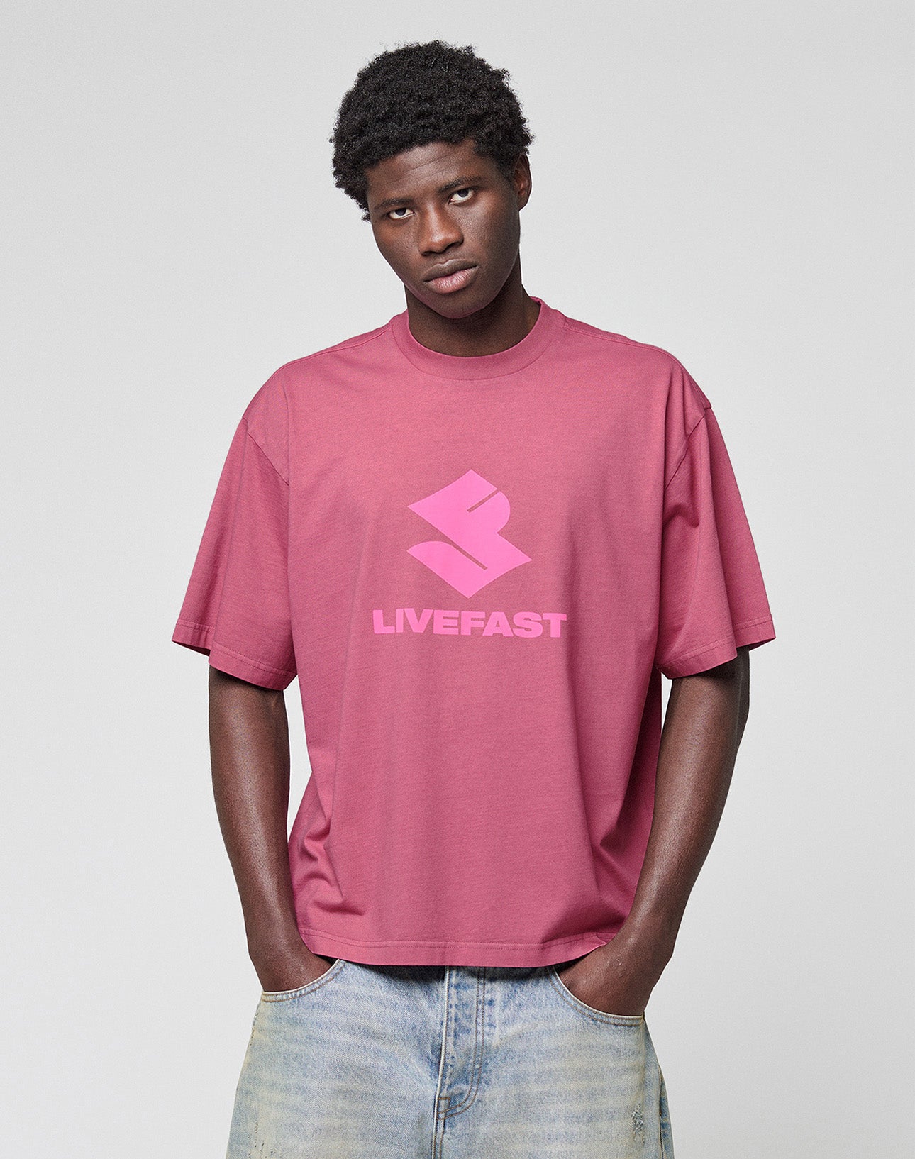 A person wearing the LIVE FAST DIE YOUNG Live Fast LF Tee, an oversized rose 100% Baumwolle T-shirt with bold LIVEFAST frontprint, stands with hands in pockets against a plain light background.