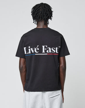 A person with short dreadlocks faces away from the camera, wearing a black LIVE FAST DIE YOUNG Live Fast Paris Tee. The Regular Fit shirt is 100% Baumwolle and features a bold Livé Fast™ Backprint with a red, white, and blue underline.
