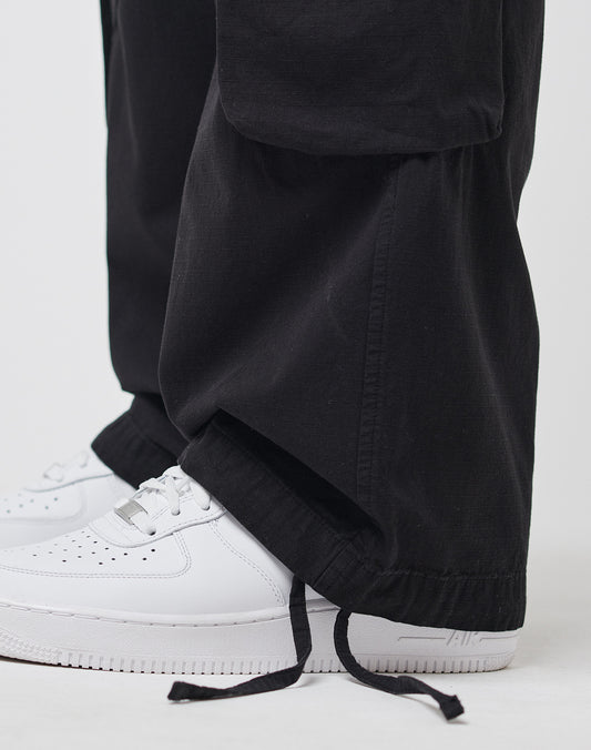 A close-up shows a person wearing LIVE FAST DIE YOUNG Wide Summer Cargo pants in black, featuring a loose fit, Cargotaschen, and drawstring hems draped over white Nike Air Force 1 sneakers against a plain white background.