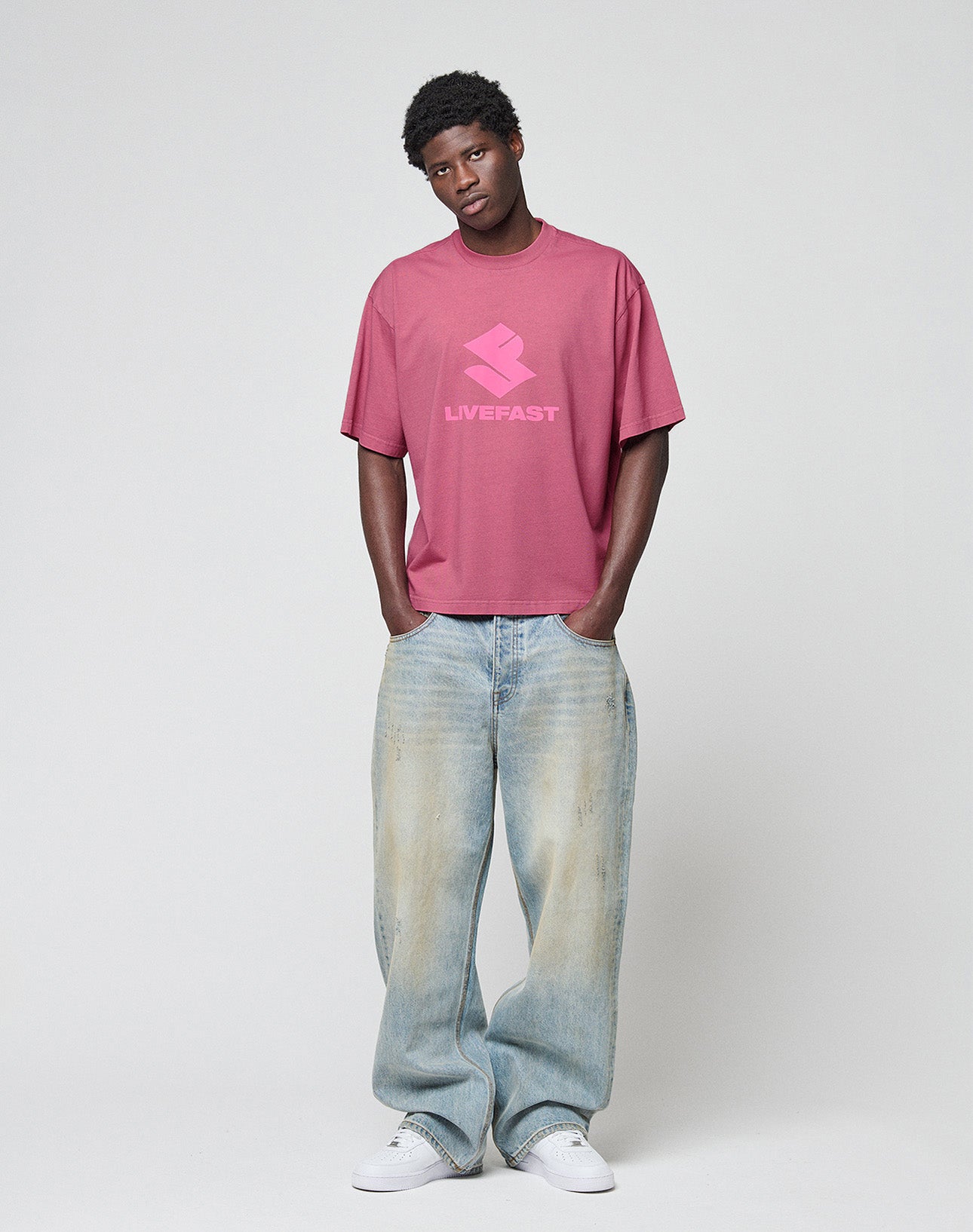 A man stands against a plain background wearing a loose pink LIVE FAST DIE YOUNG t-shirt, LFDY Baggy Denim jeans, and white sneakers, with his hands in his pockets and a relaxed expression.