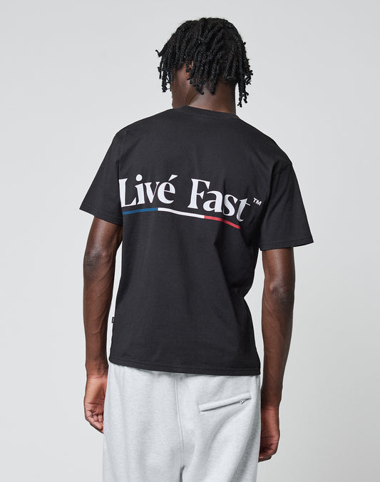 A person with short, twisted hair faces away, wearing a black Live Fast Paris Tee by LIVE FAST DIE YOUNG—100% Baumwolle, front and back print “Live Fast” in large white letters—and light gray sweatpants, against a plain gray background.