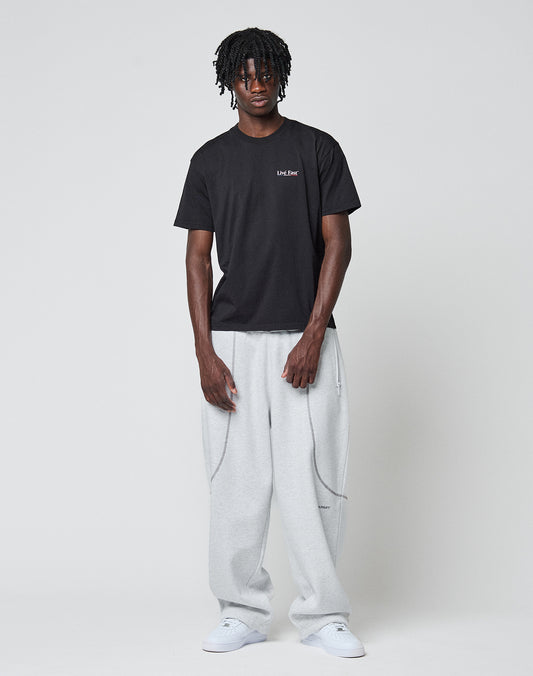 A man with short dreadlocks wears the LIVE FAST DIE YOUNG Live Fast Paris Tee with front and back print, light gray baggy sweatpants, and white sneakers, standing against a plain light gray background.