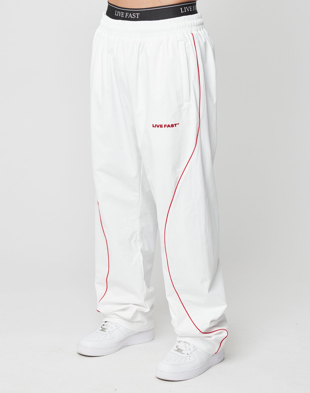 A person wears LIVE FAST DIE YOUNG LFDY Wavy Trackpants—loose white with red piping, Wide Fit, black waistband with LIVE FAST in white, and Silikonprint text on the left thigh—paired with white sneakers. Background is plain white.