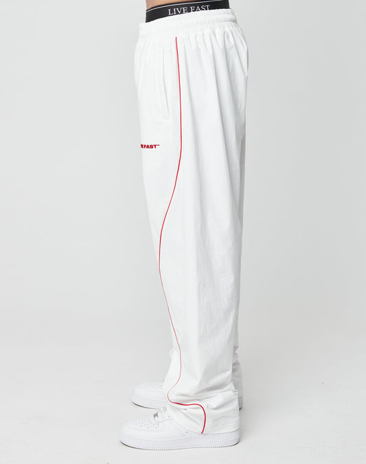 A person in LIVE FAST DIE YOUNG’s LFDY Wavy Trackpants, styled with red piping and a black Live Fast waistband, pairs them with white sneakers while standing sideways against a plain white background.