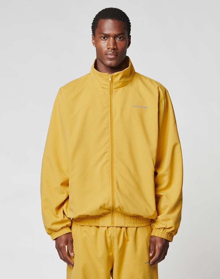 A man wears the LIVE FAST DIE YOUNG LFDY Trackjacket in an oversize fit, mustard yellow with reflective print, along with matching pants. He stands facing forward against a plain light gray background. | model