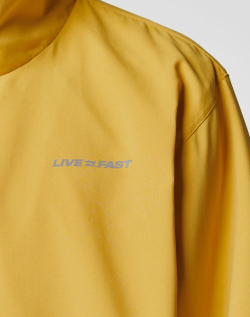 A close-up of the LFDY Trackjacket by LIVE FAST DIE YOUNG shows yellow polyester with smooth, slightly shiny material and LIVE FAST in silver reflective print on the left chest.