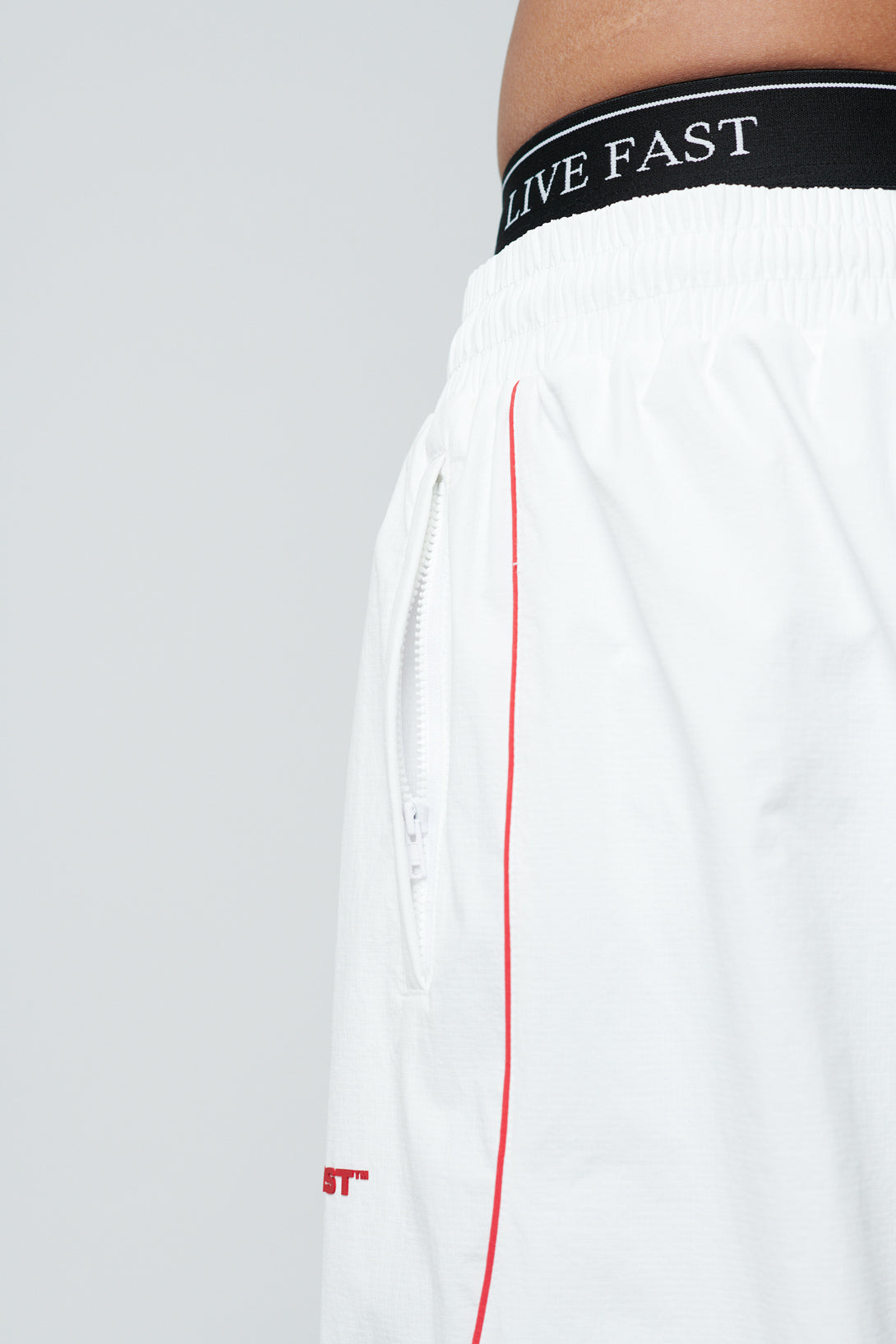 Close-up of LFDY Wavy Trackpants by LIVE FAST DIE YOUNG, featuring white fabric with red stripe, silicone print details, visible white zipper, and black elastic waistband with LIVE FAST in white text.
