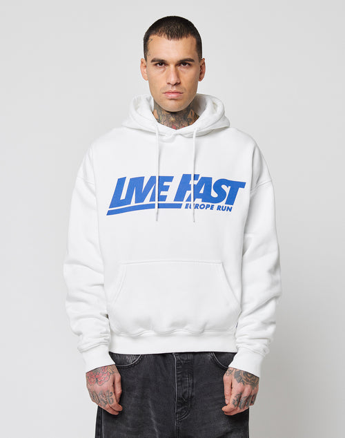 A person with short hair and visible tattoos wears the LIVE FAST DIE YOUNG Live Fast Europe Run Hooded, a white oversize hoodie with blue text, Kapuze, standing facing forward against a plain light background.