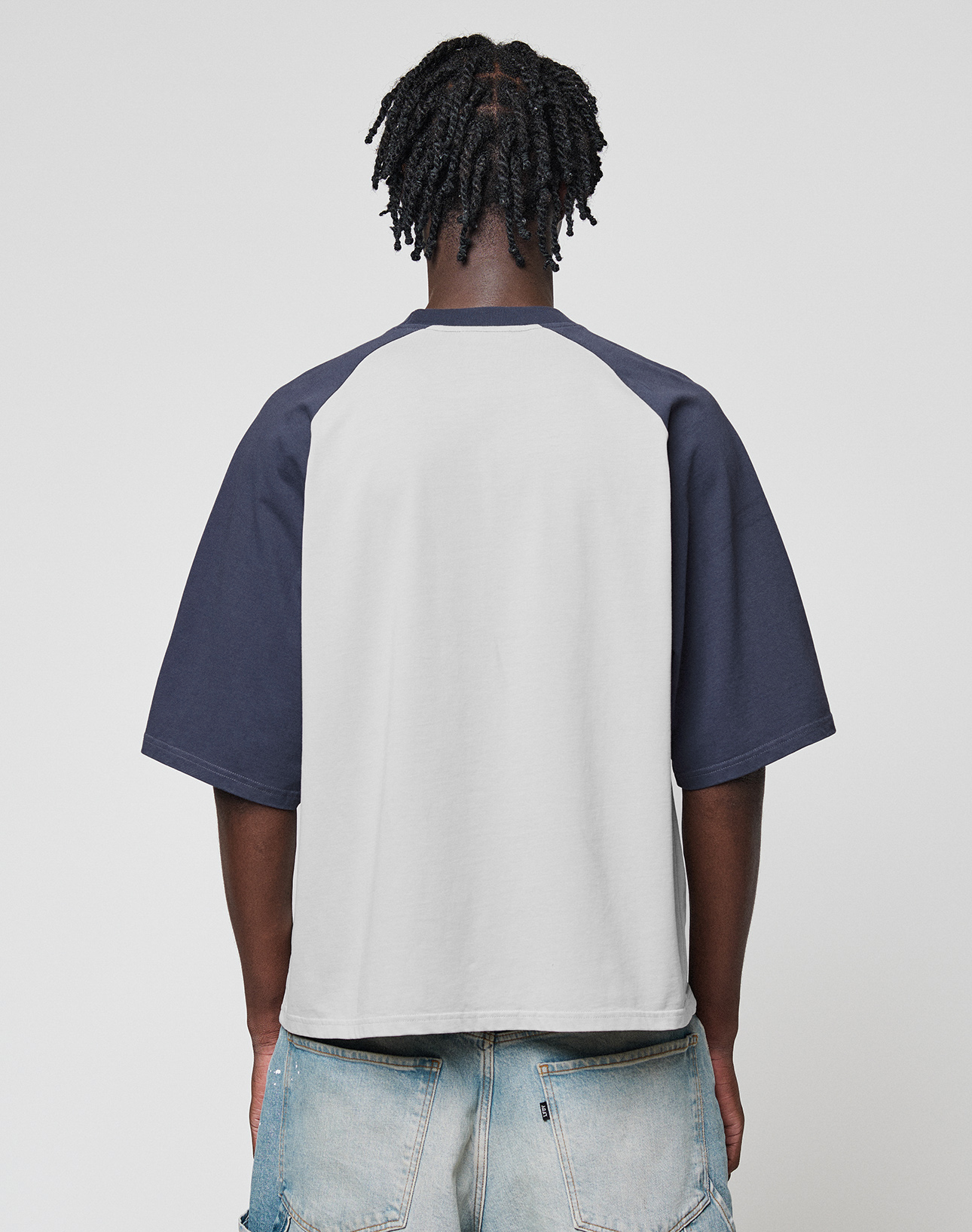 A person with short twisted hair is seen from the back, wearing the Live Fast Raglan Tee by LIVE FAST DIE YOUNG and light blue distressed jeans against a simple, light background.