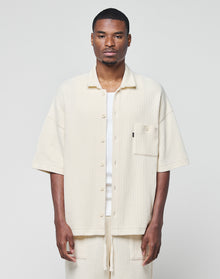 A man stands facing forward wearing the LIVE FAST DIE YOUNG Basic Knit Shirt in cream, an oversize, short-sleeve button-up with Hemdkragen and front pocket, paired with matching pants against a plain light background.