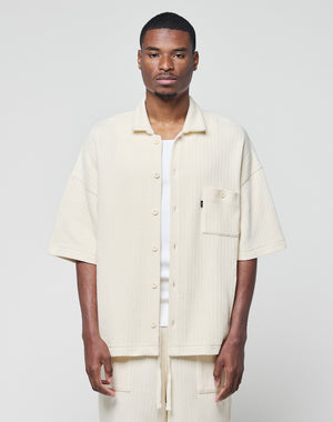 A man stands facing forward wearing the LIVE FAST DIE YOUNG Basic Knit Shirt in cream, an oversize, short-sleeve button-up with Hemdkragen and front pocket, paired with matching pants against a plain light background.