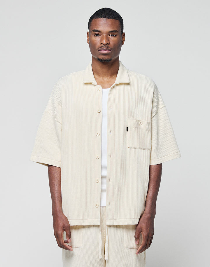 A man stands facing forward wearing the LIVE FAST DIE YOUNG Basic Knit Shirt in cream, an oversize, short-sleeve button-up with Hemdkragen and front pocket, paired with matching pants against a plain light background.