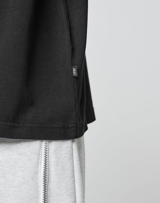 Close-up of the LFDY Paris Tee by LIVE FAST DIE YOUNG in an oversize black fit with a small side tag, paired with light gray pants featuring a visible vertical zipper. The background is plain and light-colored.